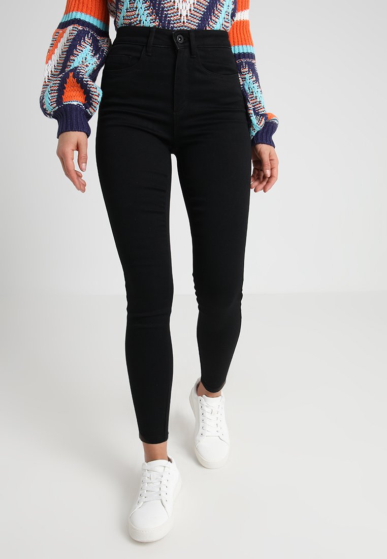 Zwarte high-waisted skinny jeans met een gladde textuur, voorzien van een knoopsluiting aan de voorkant en vier zakken, gecombineerd met witte sneakers.