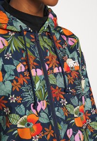 Granatowa rozpinana bluza z żywym kwiatowym wzorem, zawierająca pomarańczowe, różowe i zielone kwiaty, regulowane sznurki oraz logo na piersi.