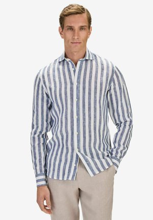 Man med blå och vit vertikalt randig långärmad skjorta och beige byxor, stående med ena handen i fickan.