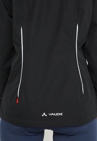 Black jacket with white side zippers, textured fabric, and a small red accent. Features a logo "VAUDE" at the lower back.