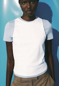 Donkerhuidige persoon in een witte mouwloze top, gedragen over een lichtblauw kortmouwshirt, staande tegen een blauwe achtergrond.