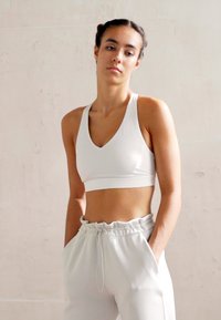 Bílá sportovní podprsenka s výstřihem do V a širokými ramínky, doplněná bílými vysokými kalhotami s elastickým pasem a bočními kapsami.