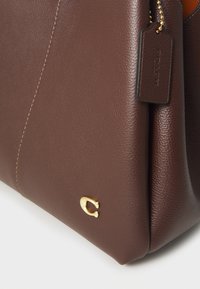 Angolo di borsa a mano in pelle marrone strutturata con logo in metallo dorato e etichetta in pelle abbinata su catena dorata.