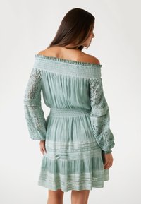 Mintgrünes schulterfreies Kleid mit langen Spitzenärmeln, gerafftem Taillenbereich und gestuften Saum. Textil mit strukturiertem Stoff und floralen sowie geometrischen Mustern.