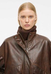 Pruun nahktagi kõrge kraega, millel on ees lukuga kinni pandav osa, nööbid ja kaks suurt taskut. Näha on kudumisega tumepruun kõrgkaelus.