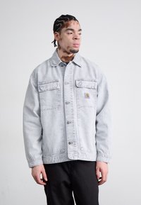 Light gray denim jacket with two front pockets, silver buttons, and subtle stitching details. Worn over a dark shirt.