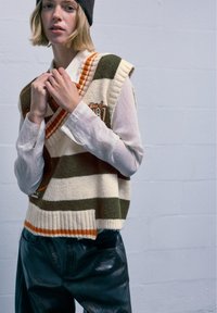 Strickloser Pullover mit Streifen in Creme, Grün und Orange, kombiniert mit einem weißen strukturierten Hemd und glänzenden schwarzen Hosen.