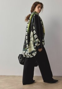 Pārāk liels melns mētelis ar kr āsainu rakstu šalli, kurā ir zaļas un krēmkrāsas akcenti. Brīvi pieguļošas melnas bikses un maza melna soma.