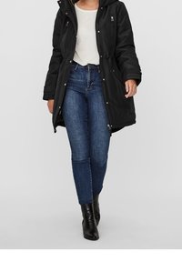Parka noire avec une capuche, des accessoires en métal argenté et des poches zippées, portée par-dessus un haut clair et associée à un jean bleu foncé et des bottines noires.