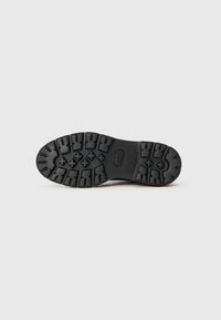 Semelle de chaussure en caoutchouc noir avec un motif de relief profond et un logo rectangulaire. Surface texturée avec de grandes crampons pour l'adhérence et la traction.