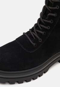 Bottine noire en suède avec surface texturée, bout rond, semelle en caoutchouc épaisse et lacets noirs passant par des œillets.