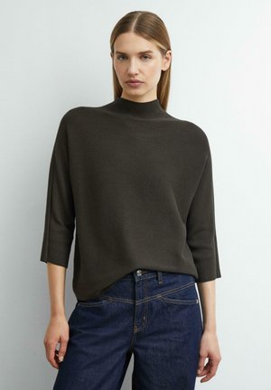 Femme aux cheveux raides portant un pull marron foncé à col montant avec des manches trois-quarts et un jean bleu sur un fond uni.