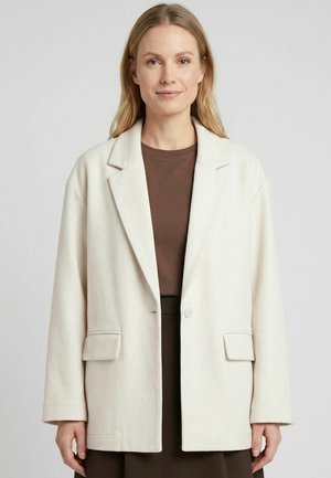 Žena s rovnými blond vlasmi nosí biely oversized blejzer cez hnedý top a sukňu, stojaca pred jednoduchým svetlým pozadím.