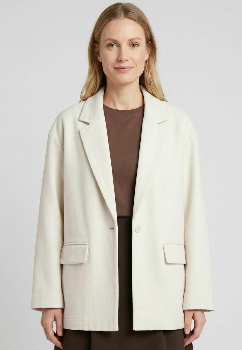 Femme aux cheveux blonds raides portant un blazer blanc oversize sur un haut marron et une jupe, debout devant un fond clair uni.