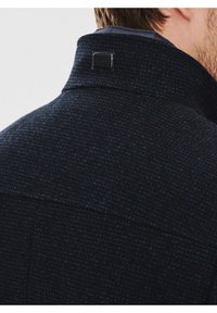 Ciemnoniebieska kurtka teksturowana z wysokim kołnierzem, z małą czarną metką logo z tyłu. Materiał sprawia wrażenie tkanej i mocnej.