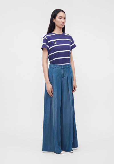Femme portant une chemise à manches courtes rayée violette et blanche avec un petit écusson, associée à un pantalon bleu large taille haute.