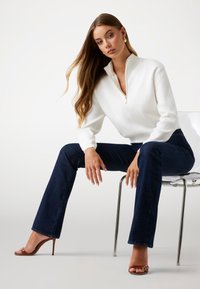 Pull blanc à fermeture éclair avec texture côtelée, associé à un jean bleu foncé évasé. Le mannequin porte des sandales à talons bruns, assis sur une chaise transparente.