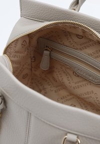 Wnętrze torby z jasnoszarej skóry, z kieszonką na zamek i wyściółką z jasnobezowych materiałów z powtarzającym się tekstem i złotymi akcentami.