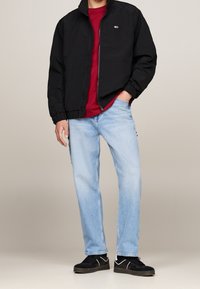 Zwarte lichtgewicht jas met elastische manchetten, rode T-shirt, lichtblauwe rechte jeans en zwarte sneakers met witte accenten.