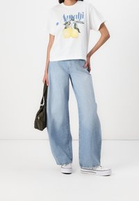 Camiseta blanca de manga corta con gráfico de limón y texto "Amalfi", combinada con jeans de pierna ancha en azul claro y zapatillas blancas. Bolsa verde oliva.