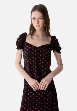 Giovane donna che indossa un vestito nero con stampa floreale rossa e maniche corte arricciate, in piedi con le mani incrociate davanti.