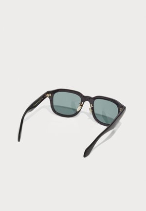 Ochelari de soare rectangulari negri cu lentile verzi, având accente aurii pe balamale și un finisaj lucios. Brațele sunt elegante și ușor curbe.