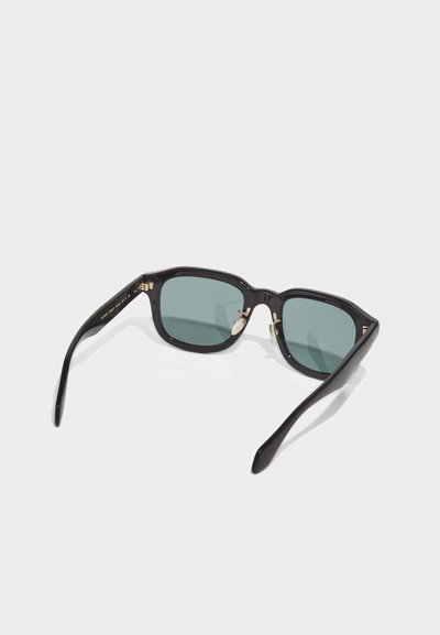 Occhiali da sole rettangolari neri con lenti scure, posati su una superficie bianca, visti da una prospettiva leggermente inclinata dall'alto.