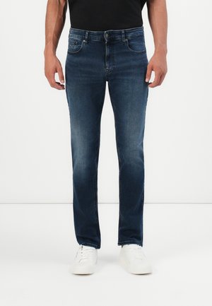 Moški v temno modrih slim-fit kavbojkah in belih supergah stoji ob običajni beli ozadju.