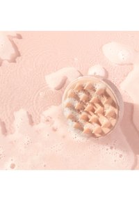 Pinkfarbener Schaum umgibt eine runde Scheuerbürste mit weichen, konisch geformten Borsten, die auf einer nassen Oberfläche ruht.