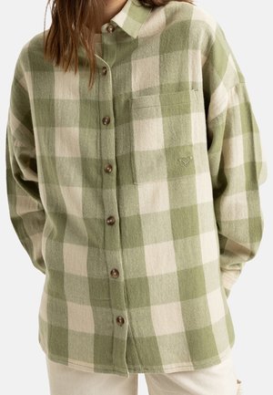 Femme portant une chemise oversize à carreaux verts et beiges avec col et poche poitrine, associée à un pantalon de couleur claire.