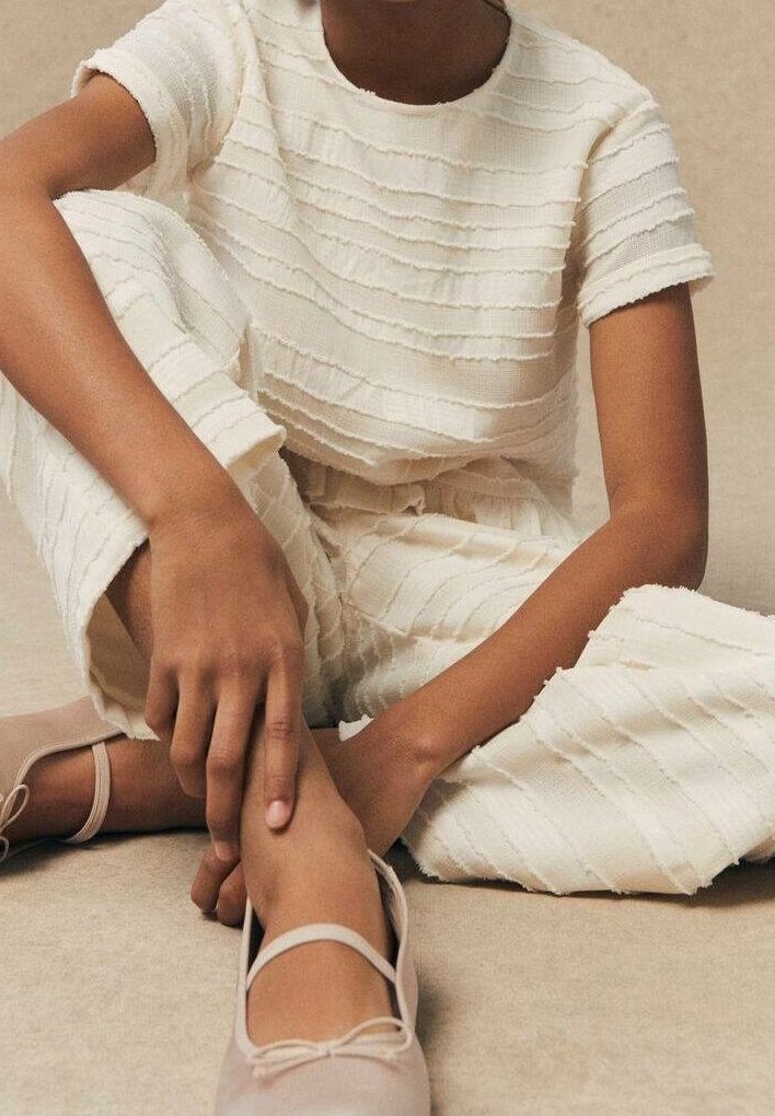 Personne portant un haut à manches courtes crème texturé et un pantalon assorti, assise en tailleur, avec des ballerines beiges et les mains reposant sur une cheville.