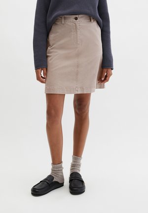 Personne portant une jupe beige mi-longue, un pull bleu marine, des chaussettes grises et des mocassins noirs, debout sur un fond blanc uni.