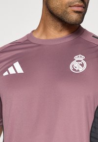 Μωβ αθλητική μπλούζα με στρογγυλή λαιμόκοψη, κοντά μανίκια, λευκό λογότυπο adidas στον ώμο και το έμβλημα της Ρεάλ Μαδρίτης στο στήθος.