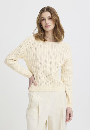 Young woman with wavy brown hair wearing a cream cable-knit sweater and loose cream trousers, standing with one hand on hip.