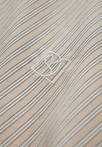 Beige fabric with vertical stripes in black and white. Features a subtle embroidered logo in white. Soft texture, medium weight.
