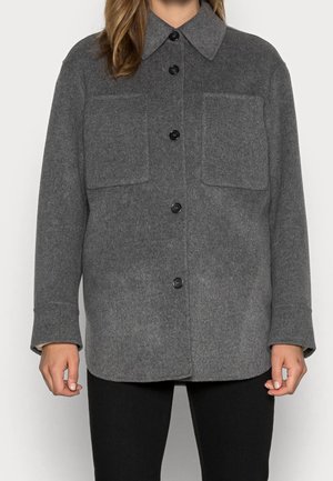 Kobieta w szarej wełnianej kurtce na guziki z dwoma przednimi kieszeniami na piersi, stojąca na jednolitym tle.