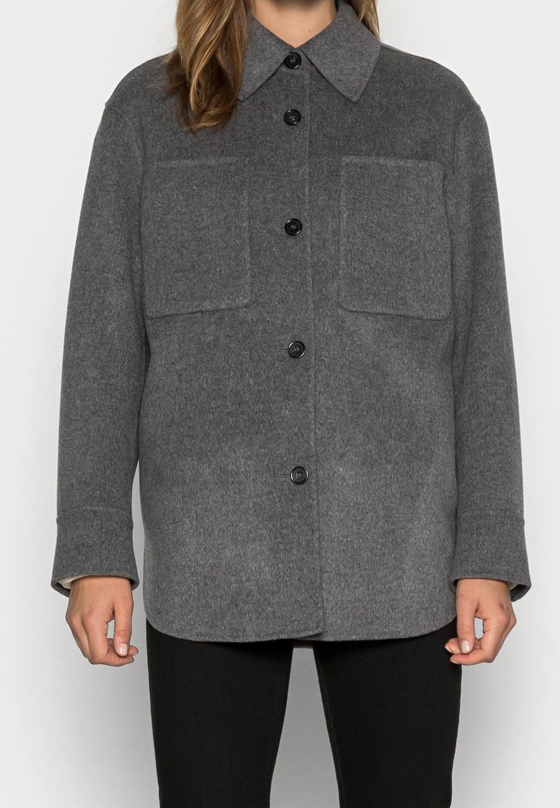 Kvinde iført en grå uldjakke med knapper og to brystlommer, stående mod en ensfarvet baggrund.