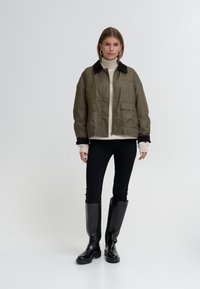 Olivgrön kort jacka med brun manchesterrygg, två framfickor, bärs över en beige tröja och svarta tajta jeans med knäslitsar.