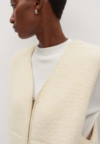 Cremefarbener Fleece-Weste mit einem glatten Reißverschluss und einer weichen, strukturierten Oberfläche. Getragen über einem weißen gerippten Rollkragenpullover. Keine Kapuze oder Ärmel.