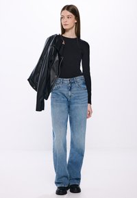 Schwarzes Langarmshirt kombiniert mit hellblauen, weit geschnittenen Jeans. Eine schwarze Lederjacke hängt über einer Schulter. Schwarze klobige Stiefel runden den Look ab.