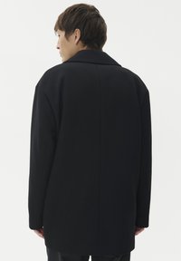 Giovane con capelli castani corti visto di spalle, indossa un cappotto nero morbido su uno sfondo chiaro e semplice.