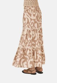 Falda larga y con volantes en beige con un ligero estampado, que cuenta con una cintura fruncida y una tela suave. Combinada con sandalias planas marrones.