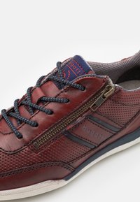 Sneaker rossa in pelle con textura perforata, lacci blu, cerniera laterale e dettagli di branding. Progettata per comfort e utilizzo informale.