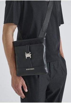 Bolso cruzado de cuero negro con herrajes plateados, que presenta un cierre de cinta en la parte delantera y un logo en relieve cerca de la parte inferior, exhibiendo un diseño elegante y moderno.