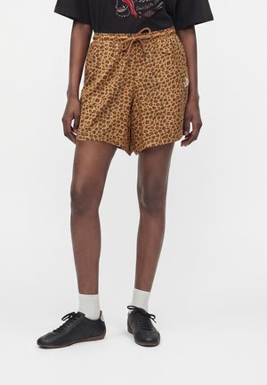 Person trägt braune Shorts mit Leopardenmuster und Kordelzug, schwarze Sneakers, weiße Socken und ein schwarzes T-Shirt mit Grafik.