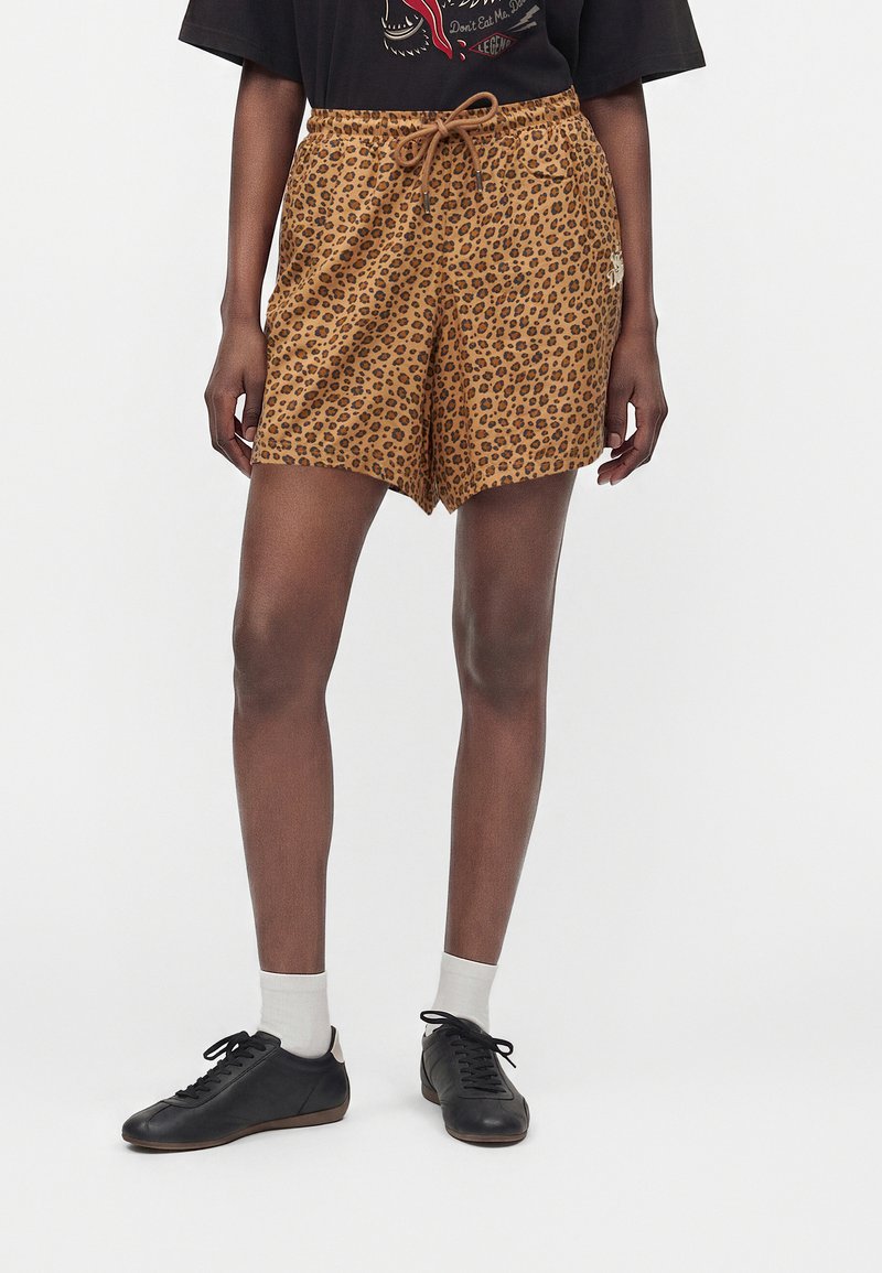 Personne portant un short marron à imprimé léopard avec cordon, des baskets noires, des chaussettes blanches et un t-shirt noir avec un motif graphique.