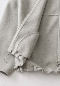 Hellgraue Wolljacke mit Reißverschluss, elastischem Bund und umgeschlagenem Ärmeldetail vor weißem Hintergrund.