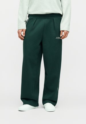 Person iført mørkegrønne viddebukser med "Le Glazik"-logo på låret, kombineret med hvide sneakers og en lysegrå langærmet top.