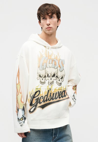 Noormees, kes kannab valget kapuutsiga dressipluusi, millel on leegitsevad koljud ja "Gcdswear" tekst, ning siniseid teksaseid, vaadates veidi kõrvale.