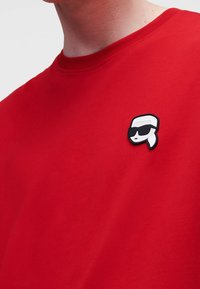 Rød bomulds T-shirt med et hvidt og sort cartoon-logo med solbriller på brystet. Kortærmet og med rund hals.